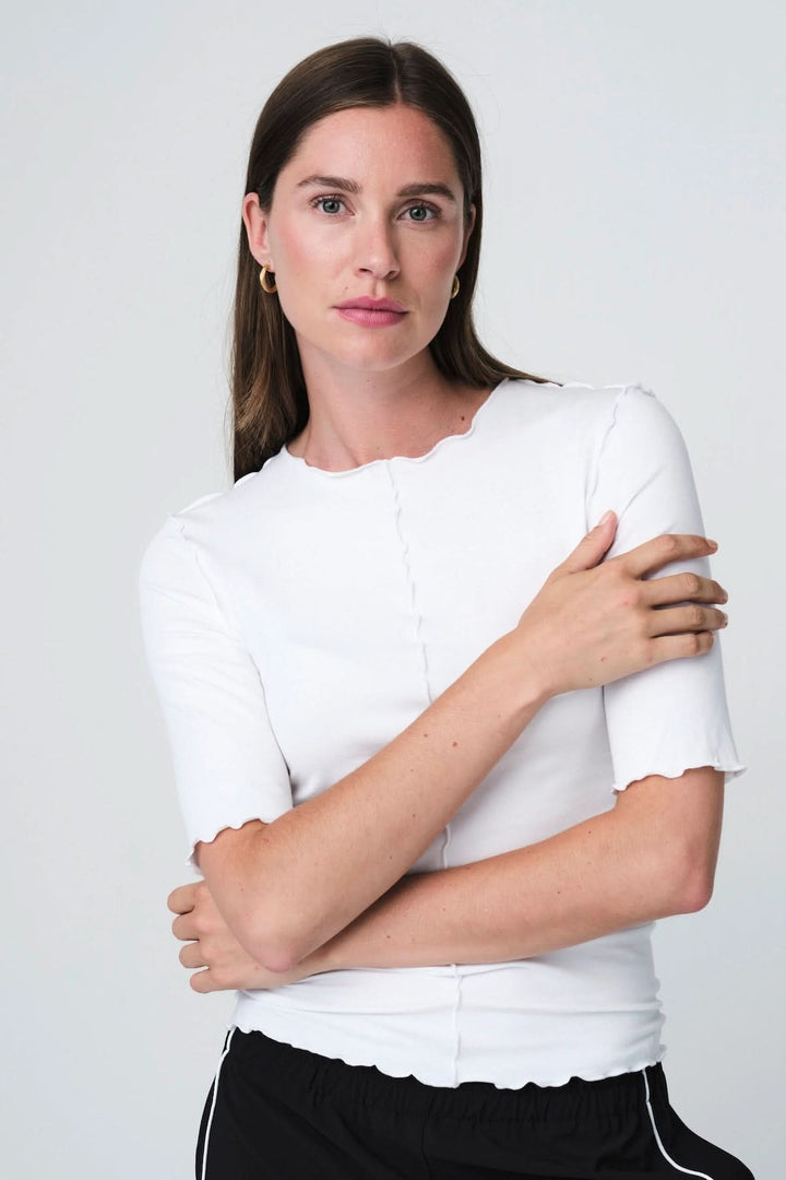 Femme portant un T-shirt ajusté à finitions ondulées blanc, manches courtes et col rond, design épuré et moderne.