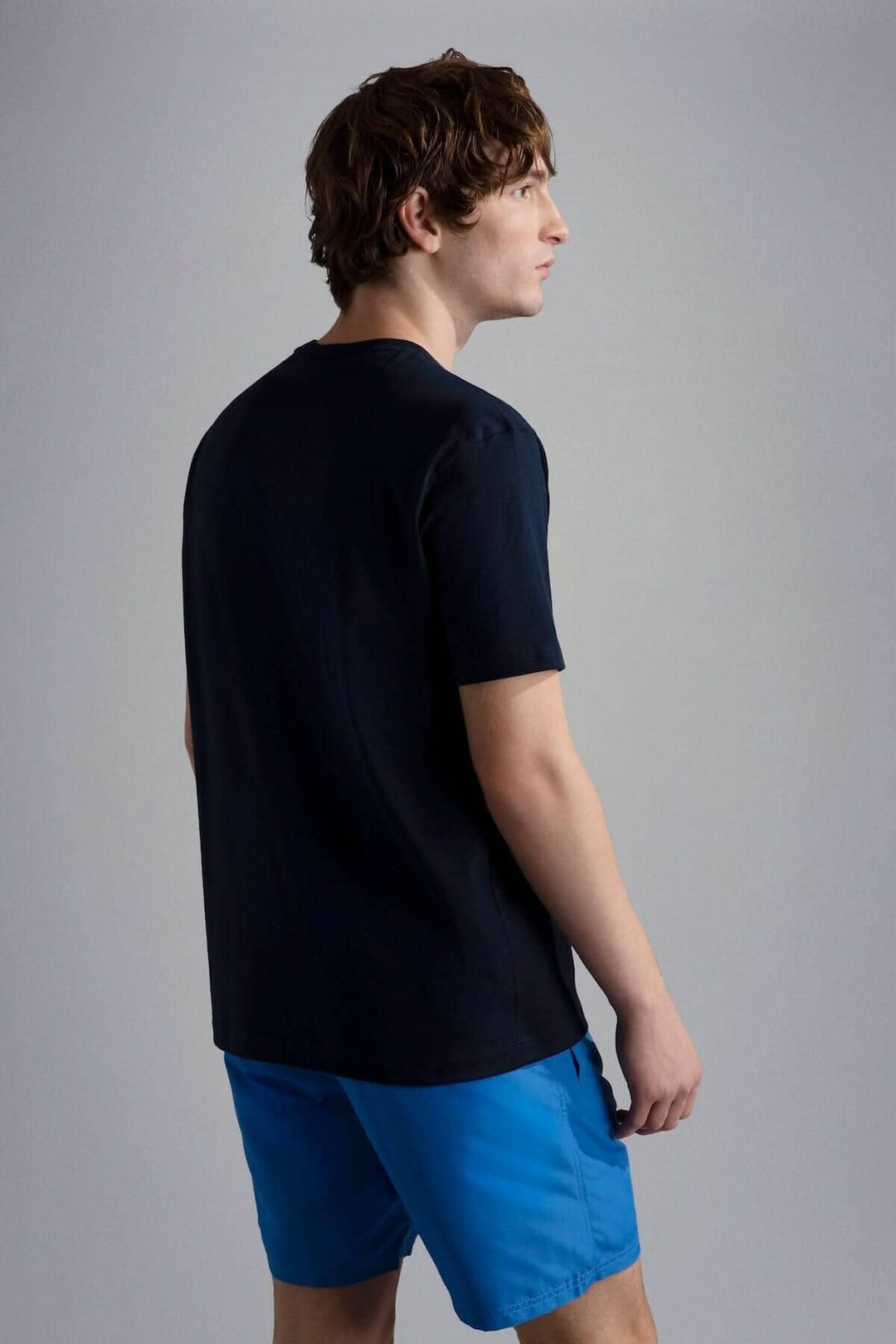 Homme portant un t-shirt noir et un short bleu, vu de dos, dans un environnement sobre.