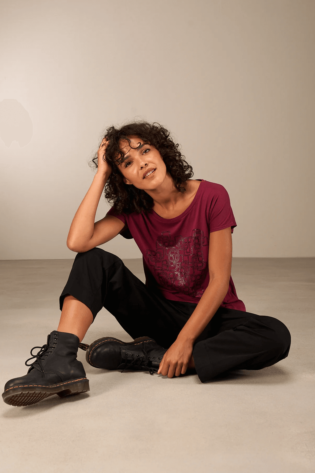 Femme posant en t-shirt imprimé graphique bordeaux, manches courtes, coupe décontractée, col rond, avec pantalon noir et bottes.