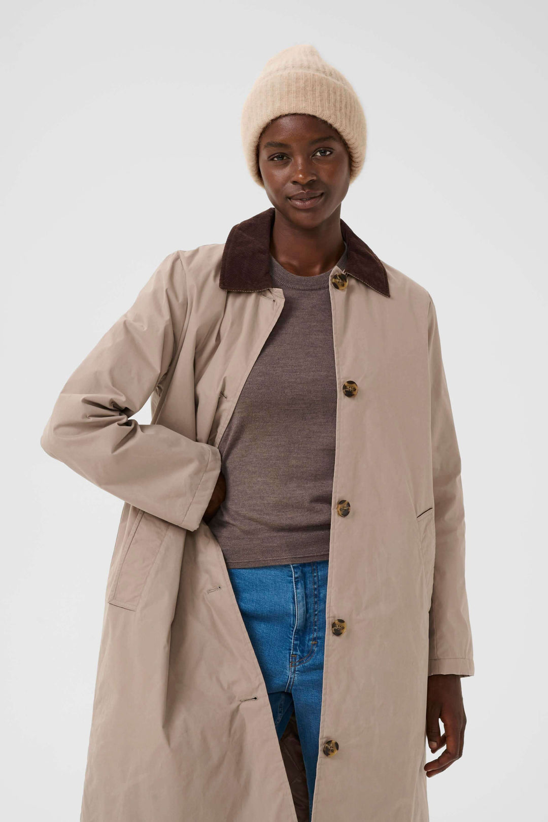 Person wearing a beige trench coat over a brown shirt with blue jeans and a beige ribbed knit beanie, displaying a casual winter style.