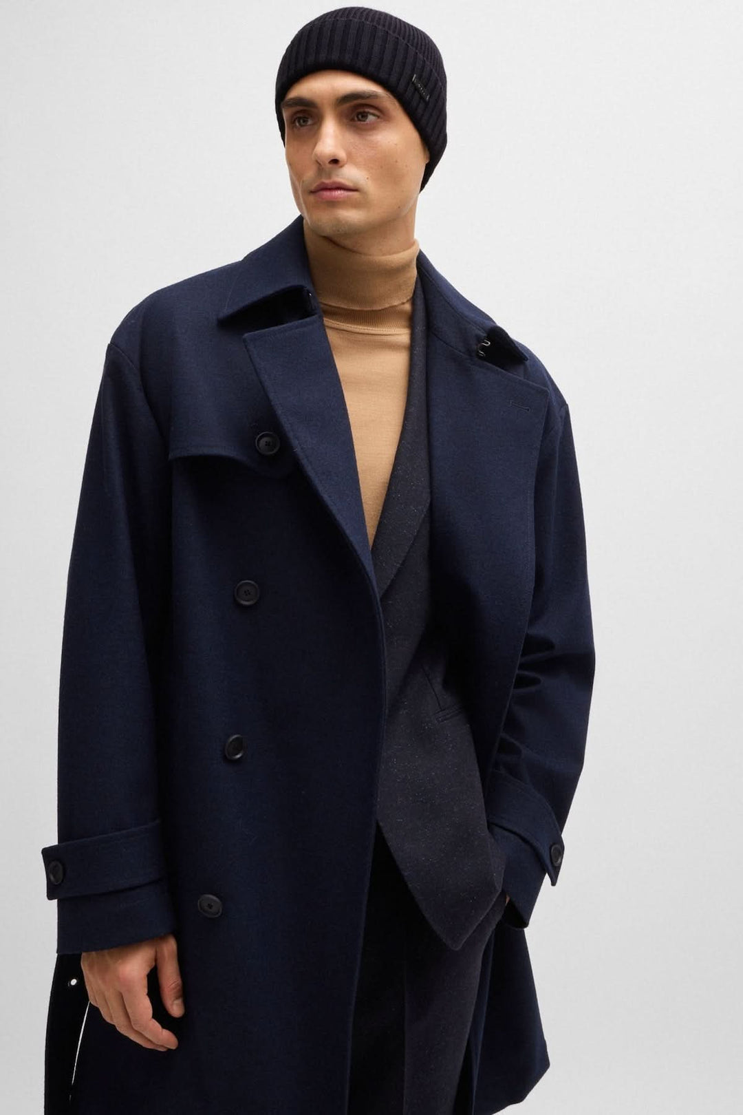 Homme portant un bonnet noir, manteau bleu marine et pull beige, dans un style élégant moderne.