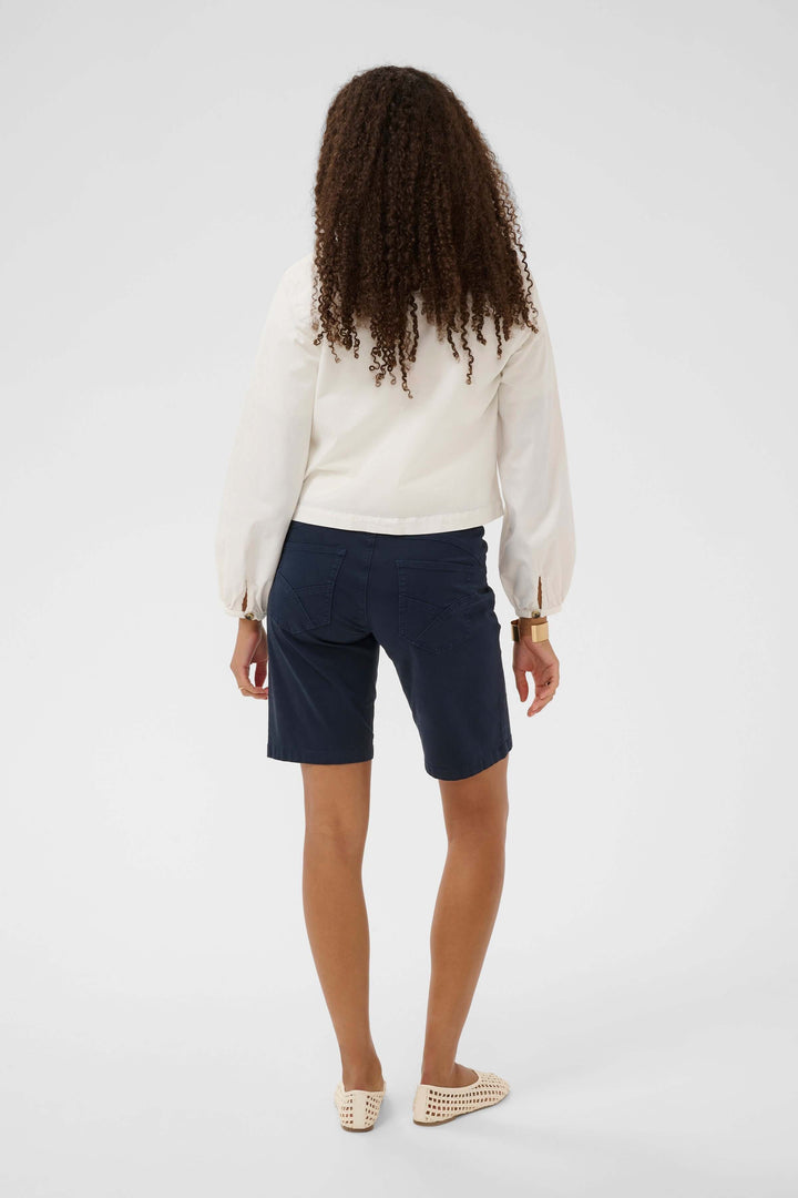Femme en short bleu marine et chemisier blanc, vue de dos, sur fond gris neutre, montrant un style chic et décontracté.