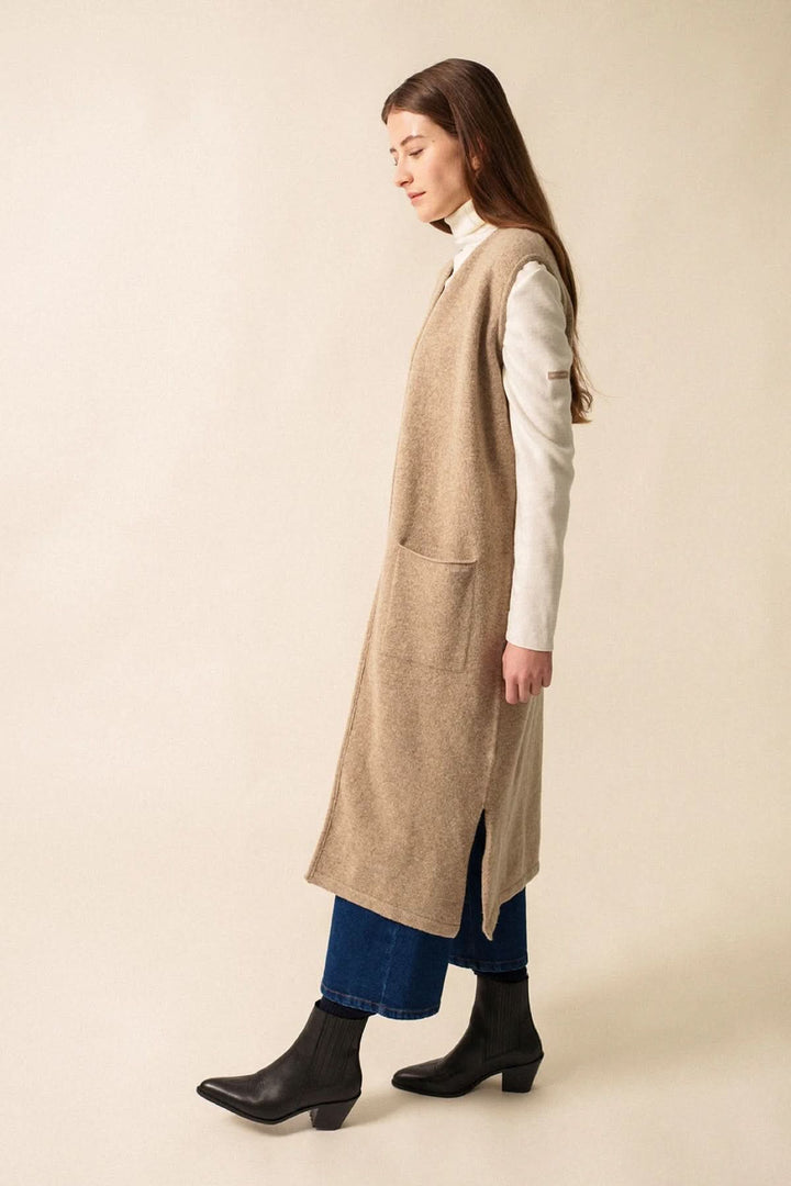 Veste longue en maille beige, sans manches, portée sur un col roulé et jean, avec poches et coupe droite.