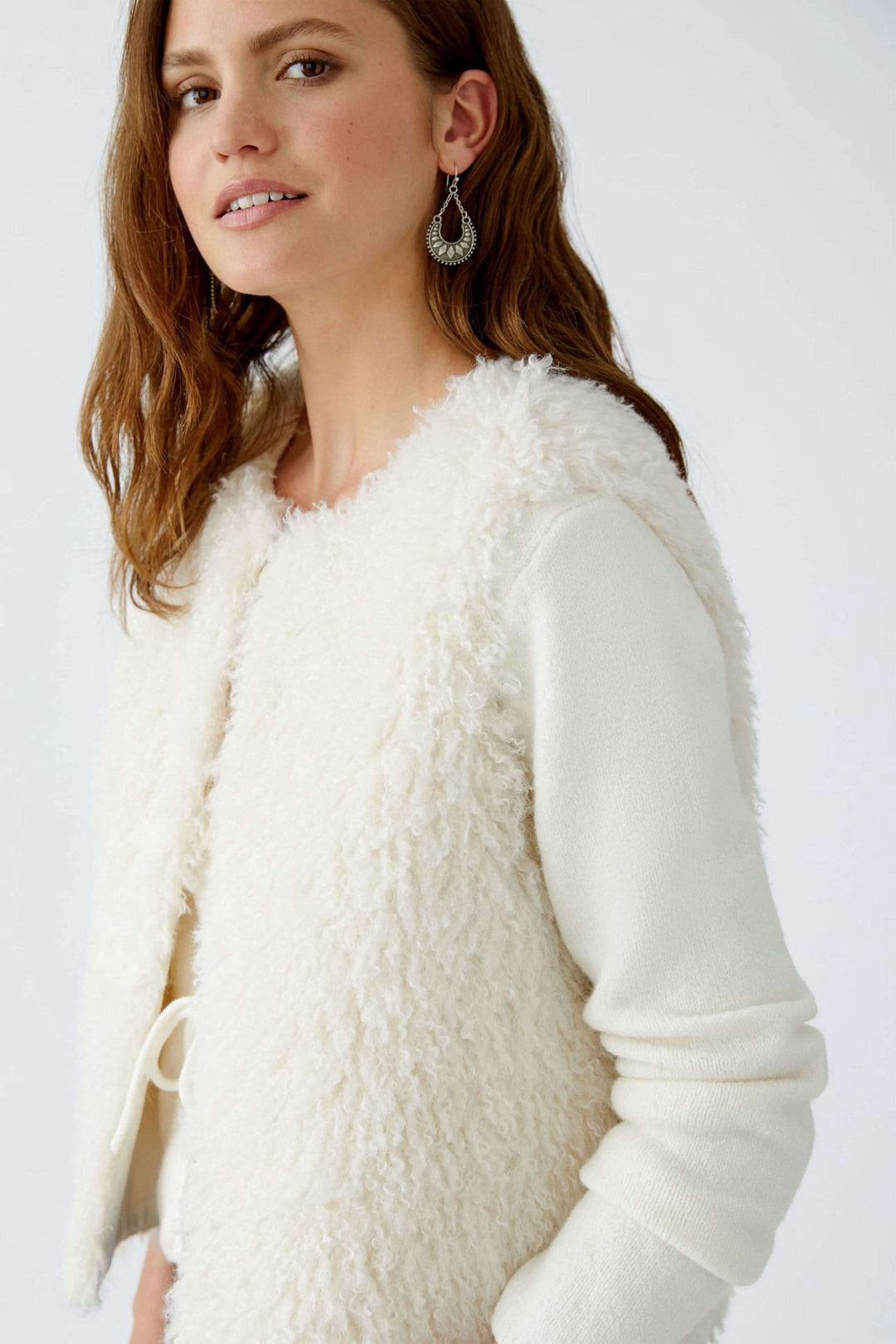 Modèle portant une veste sans manches en fausse fourrure blanche, avec des boucles d'oreilles et un look tendance.