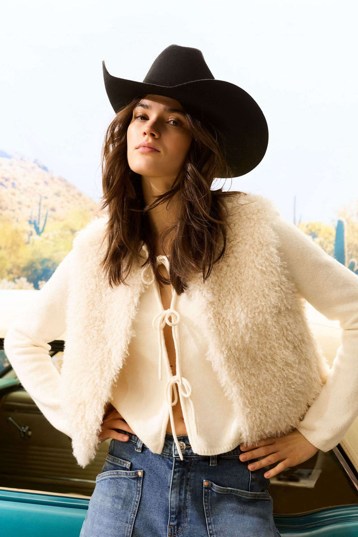 Femme portant une veste sans manches en fausse fourrure et un chapeau de cowboy, devant une voiture classique.