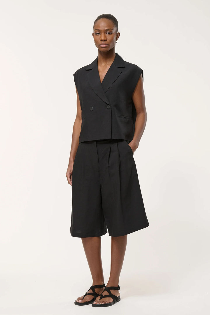 Veste tailleur Loric sans manches noire sur mannequin, coupe droite, fermée par boutons croisés, apportant une touche moderne et sophistiquée.