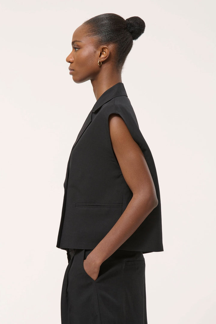 Veste tailleur Loric sans manches noire moderne avec coupe droite et col à revers, portée par un mannequin de profil pour un look élégant.