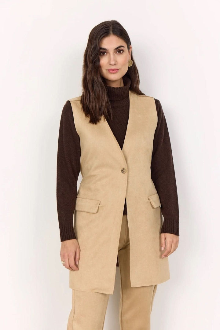 Gilet sans manches beige en matière suédée, porté sur col roulé marron, avec poches à rabat pour un look automnal.
