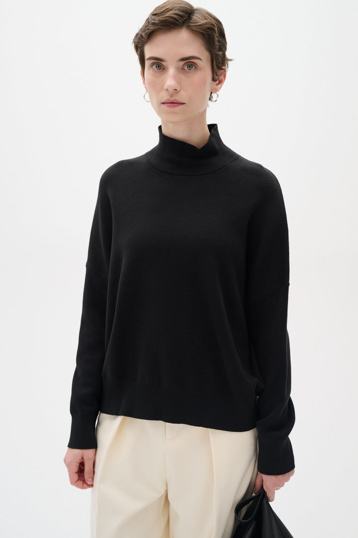 Chandail en coton noir à col roulé, manches longues, coupe ample et stylée, porté avec un pantalon crème.