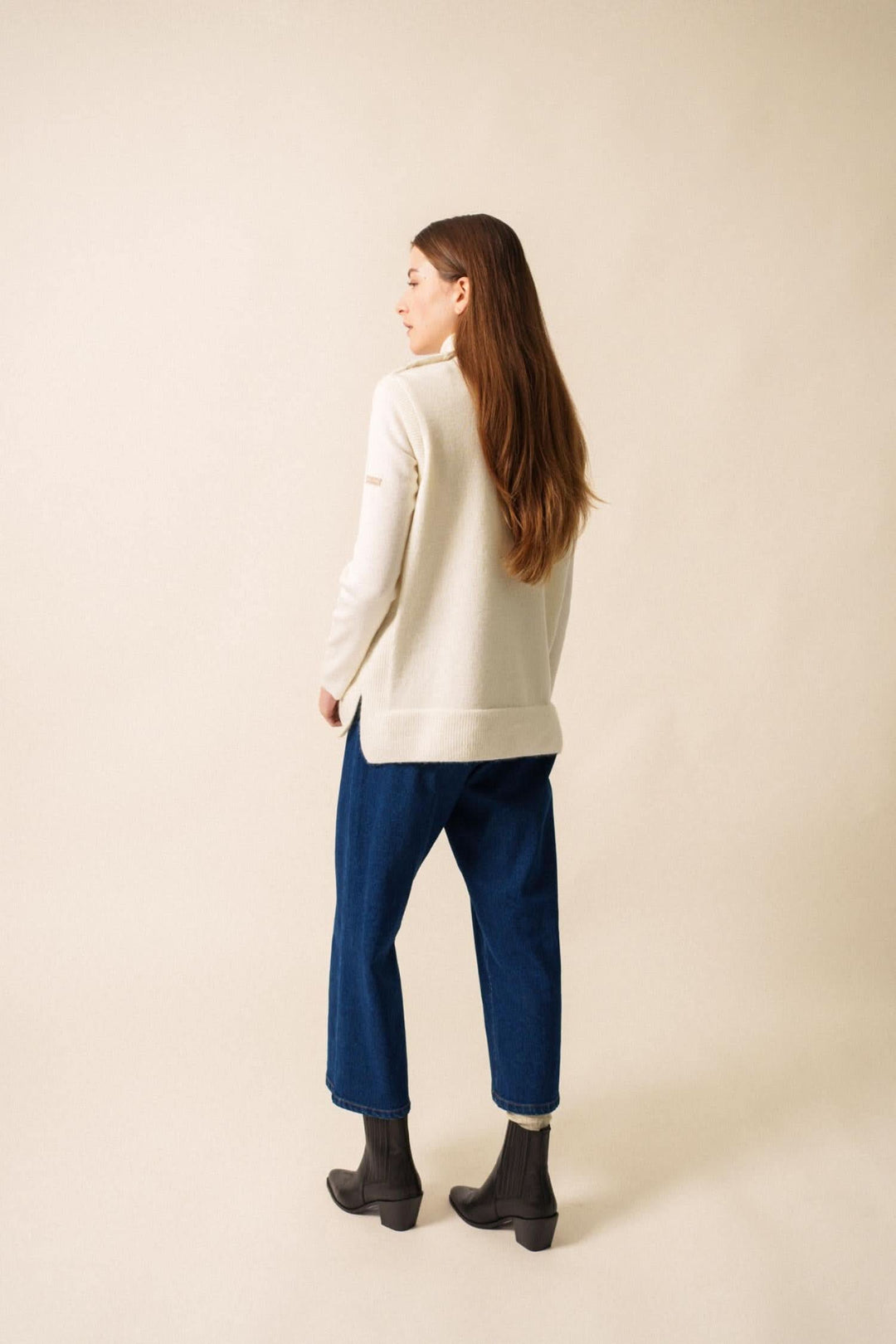 Femme de dos portant un pull crème et un pantalon bleu, style décontracté avec des bottines noires.