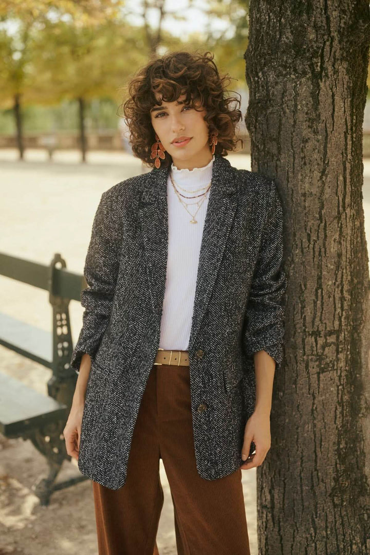 Person wearing a tweed blazer with a white t-shirt and brown trousers standing by a tree in a park setting.