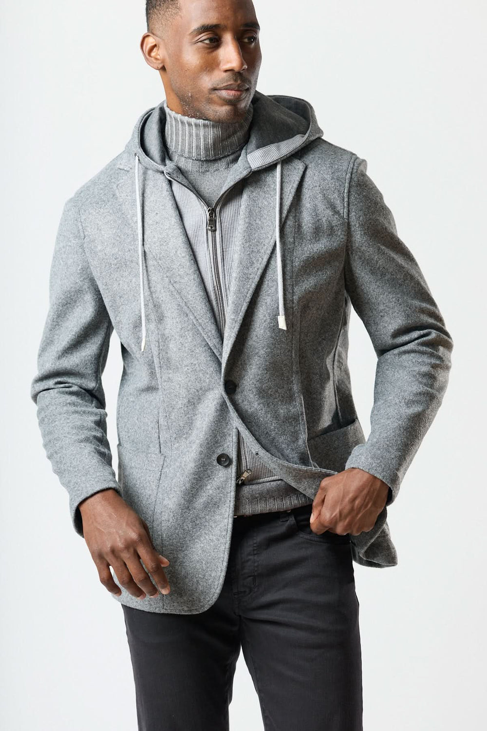 Homme portant un veston hybride à capuche gris sur un col roulé, combinant style blazer et confort, avec capuche intégrée à cordons.