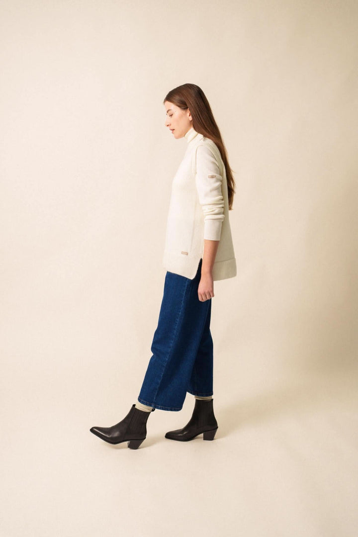 Femme portant une veste LA CLUSAZ blanche avec un jean bleu et des bottines noires, marchant sur un fond neutre.