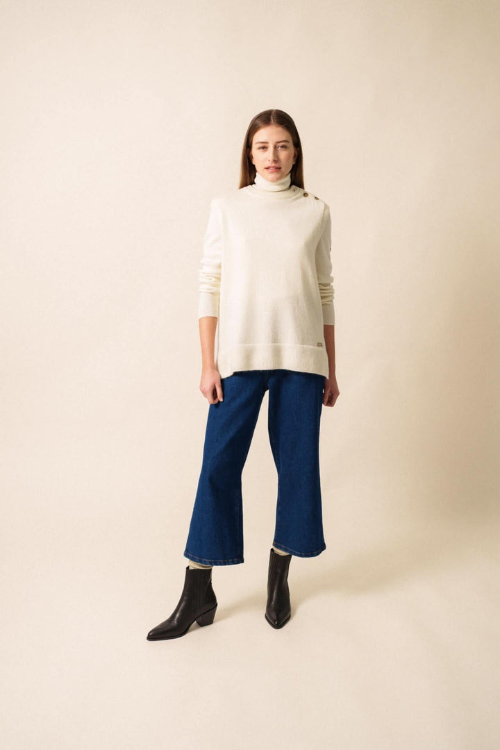 Jeune femme portant un pull blanc et un pantalon bleu, avec des bottines noires, sur fond neutre.
