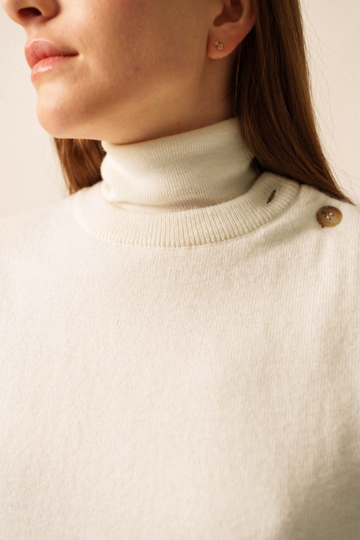 Détail d'une femme portant un pull beige à col roulé avec bouton, mettant en avant une esthétique moderne et élégante.