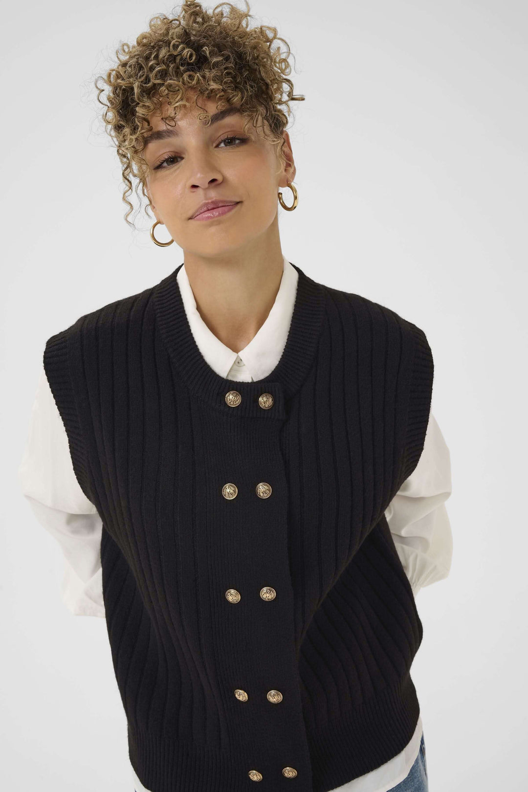 Veste Eroka sans manches en tricot à boutons dorés chic et polyvalente sur une chemise blanche, élégance décontractée.