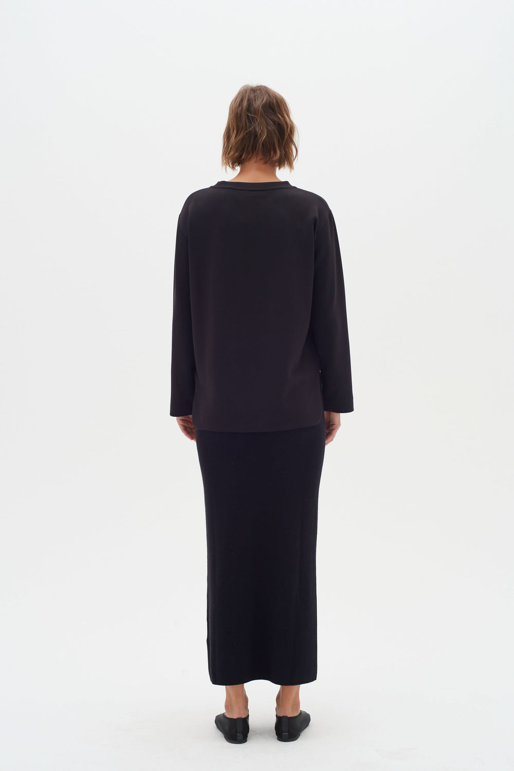 Vue arrière d'une femme portant un chandail minimaliste noir et une jupe assortie sur fond blanc.