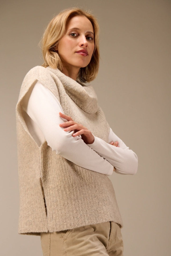 Femme portant un gilet sans manches en maille bouclée à col drapé, superposé sur un t-shirt à manches longues blanc, look élégant.