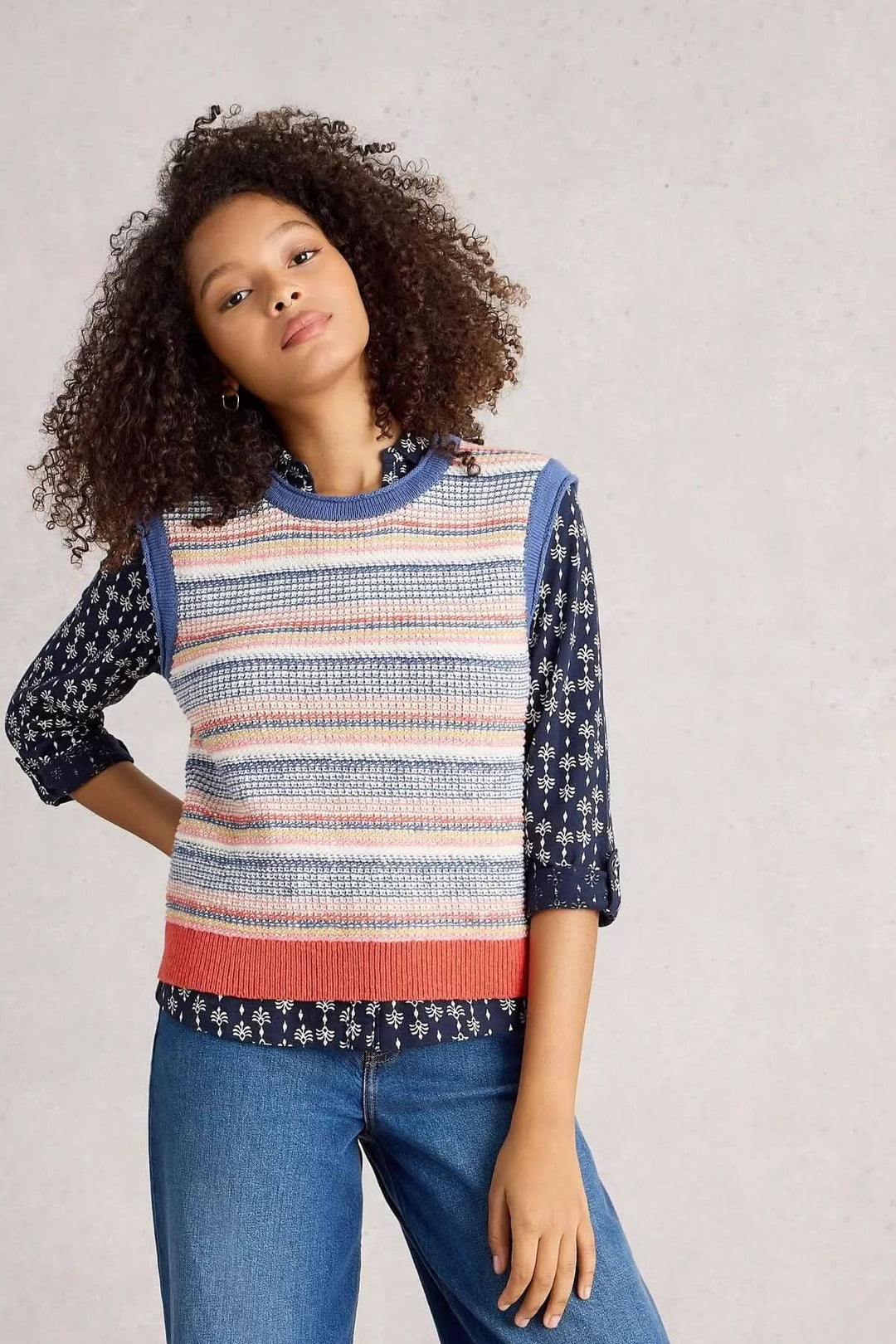 Veste sans manche Tara tricotée avec rayures multicolores, portée sur une chemise imprimée pour un look moderne et tendance.