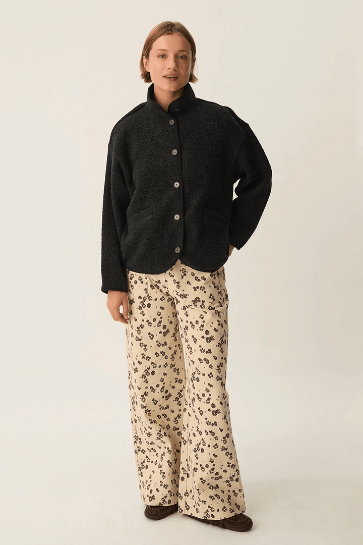 Veste Zlavina boutonnée en laine bouclée noire avec col montant, portée avec un pantalon fluide imprimé floral pour un style moderne.