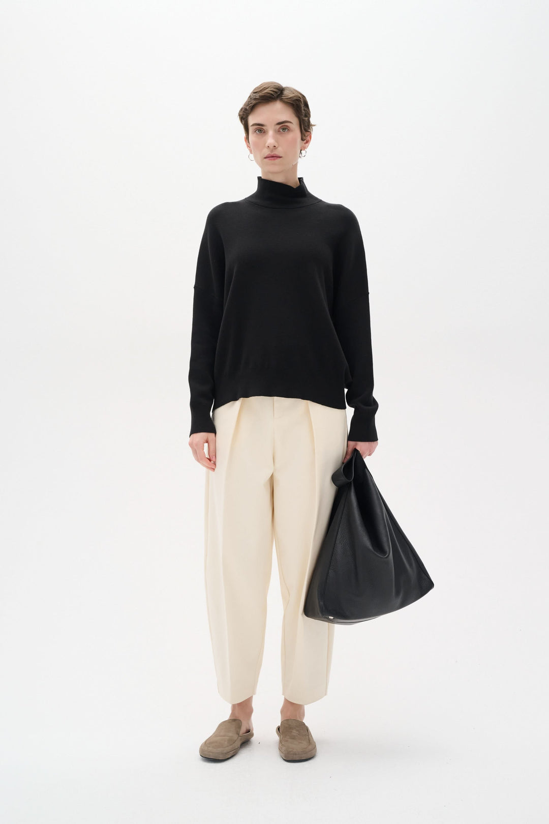 Femme en chandail noir col roulé et pantalon beige, tenant un sac noir, style décontracté et élégant.