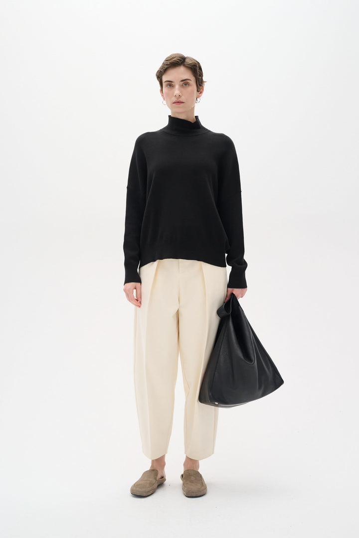 Femme en chandail noir col roulé et pantalon beige, tenant un sac noir, style décontracté et élégant.